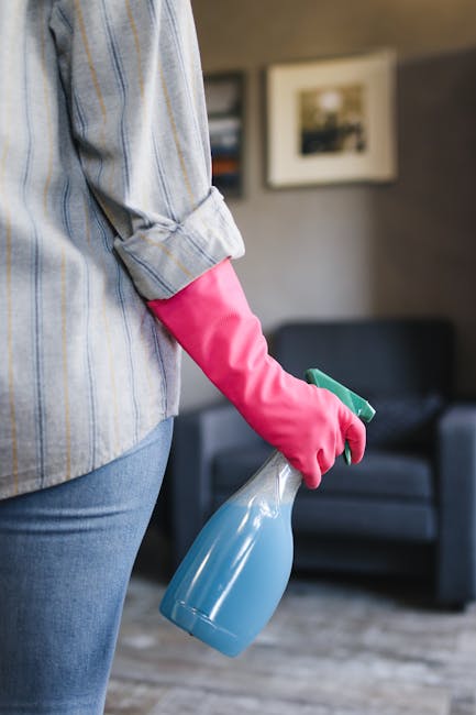 A person wearing a black jacket is using a vacuum cleaner to deep clean a yellow cushion with a white geometric pattern, placed on a dark fabric sofa in a bright room. The scene highlights surface cle