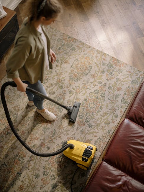A professional cleaner dressed in full protective gear, including a white coverall, face mask, and blue gloves, is performing a steam cleaning process on a plush beige carpet in a bright, modern livin